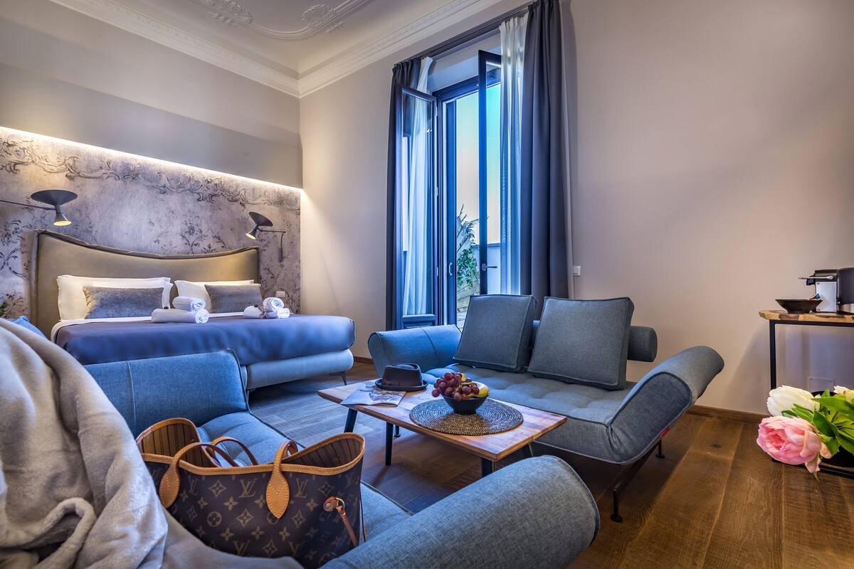 Een moderne hotelkamer met een bed, twee banken, een tafeltje met een schaal fruit en uitzicht op het omliggende landschap.