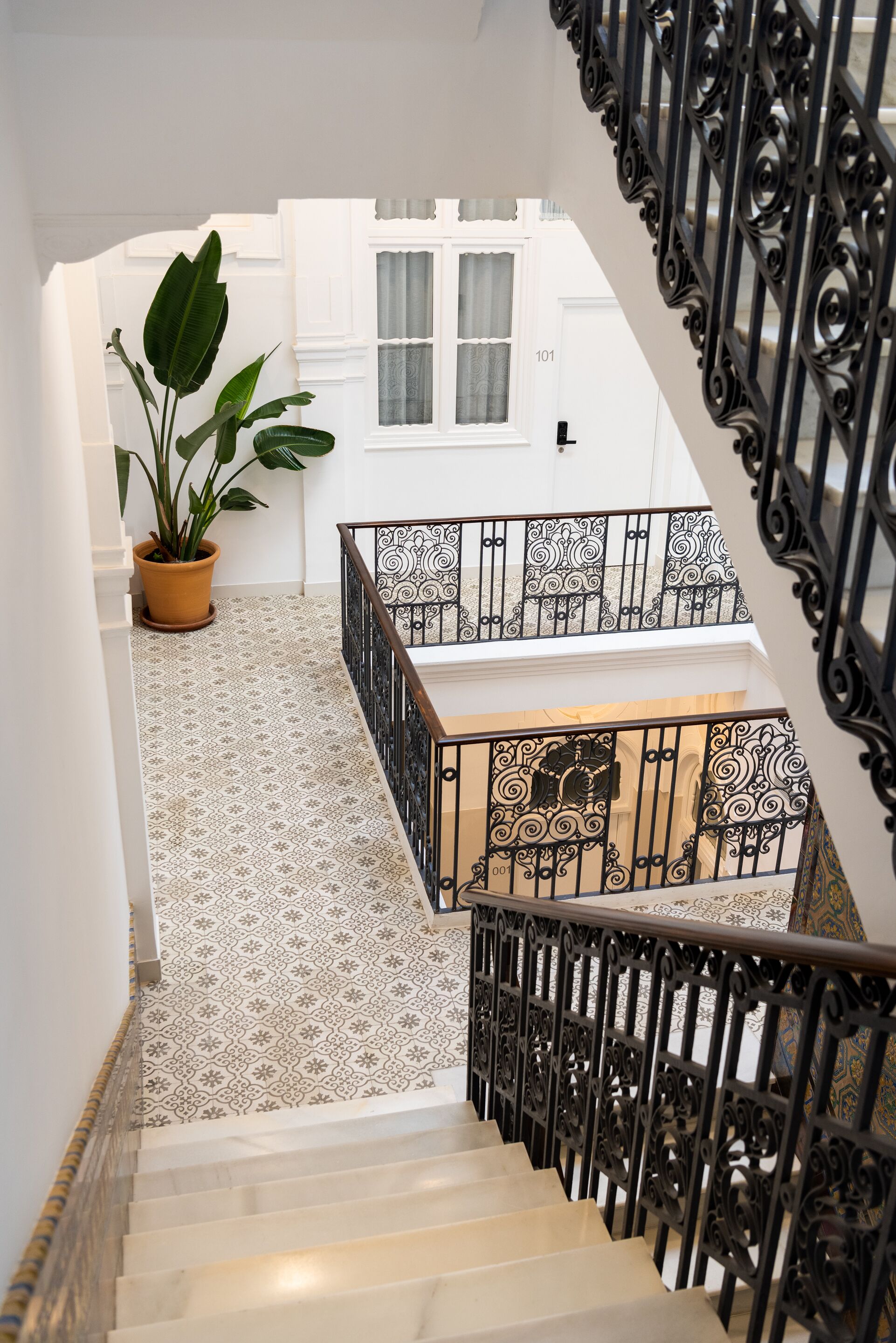 Una escalera con barandillas negras ornamentales, una planta en maceta y baldosas de suelo con diseños.
