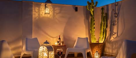 A rooftop terrace with white plastic chairs, a small table with a wine glass, a potted cactus, and a wall-mounted shower.