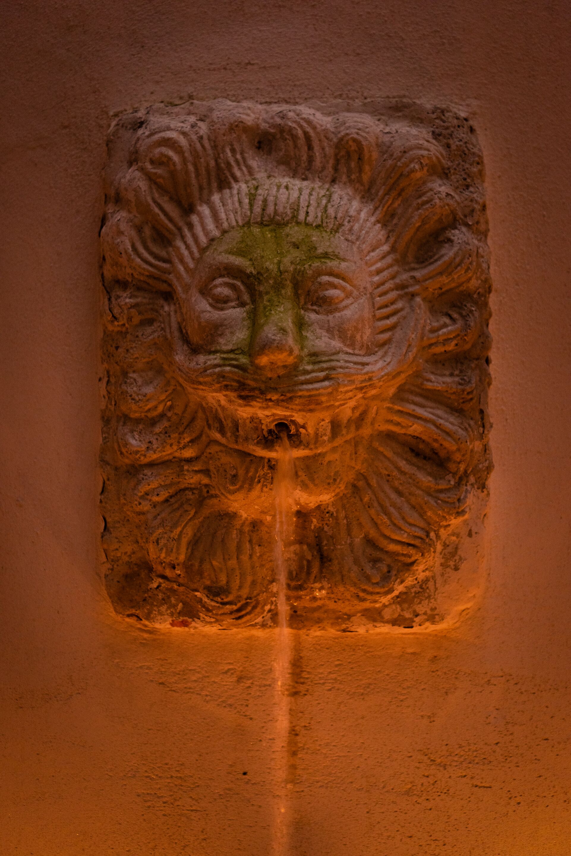 Une sculpture en pierre représentant le visage d’un lion, avec un œil et une bouche au centre, placée sur un mur sobre.