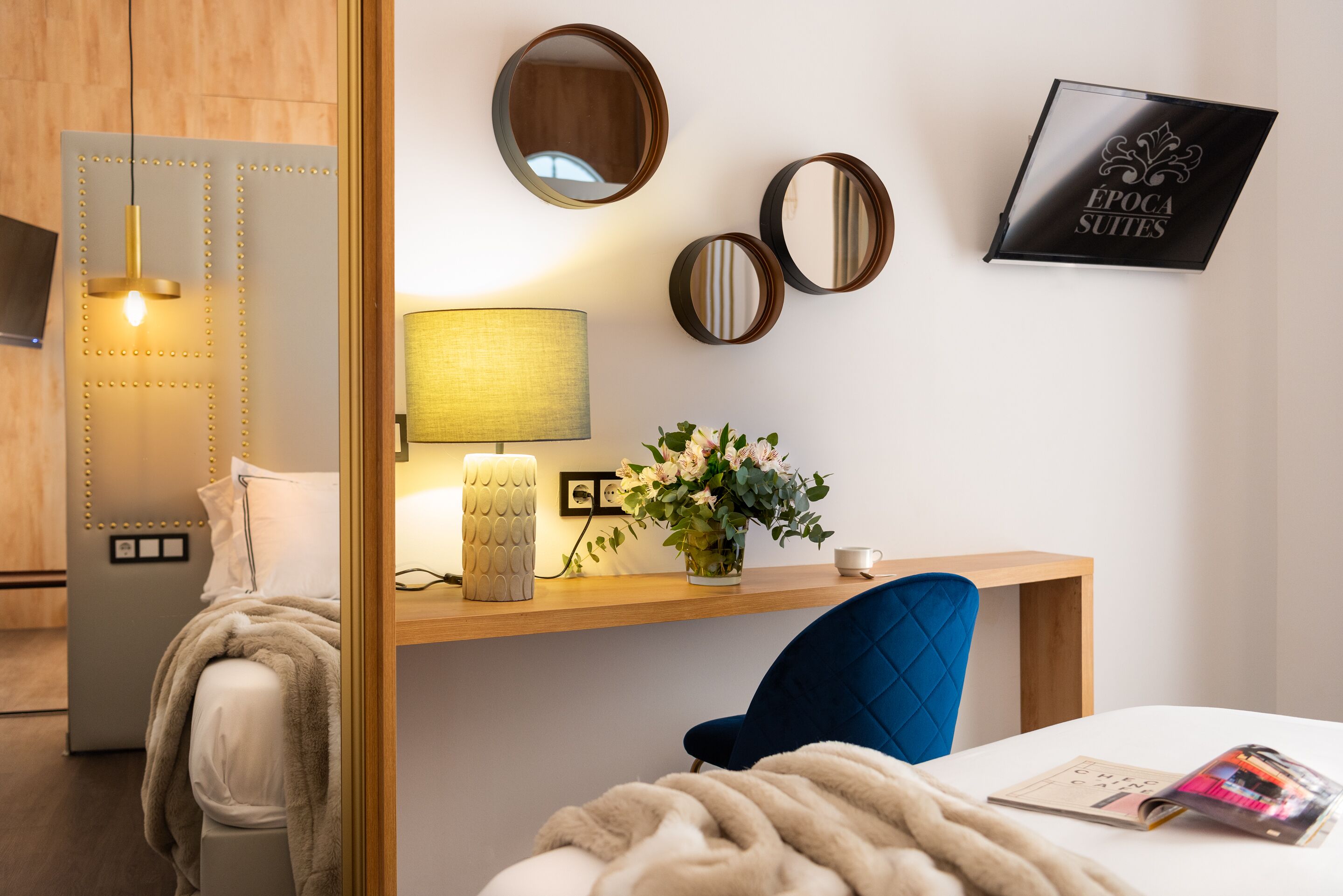 A hotel room with a bed, a desk with a lamp, a chair, and a wall-mounted mirror.