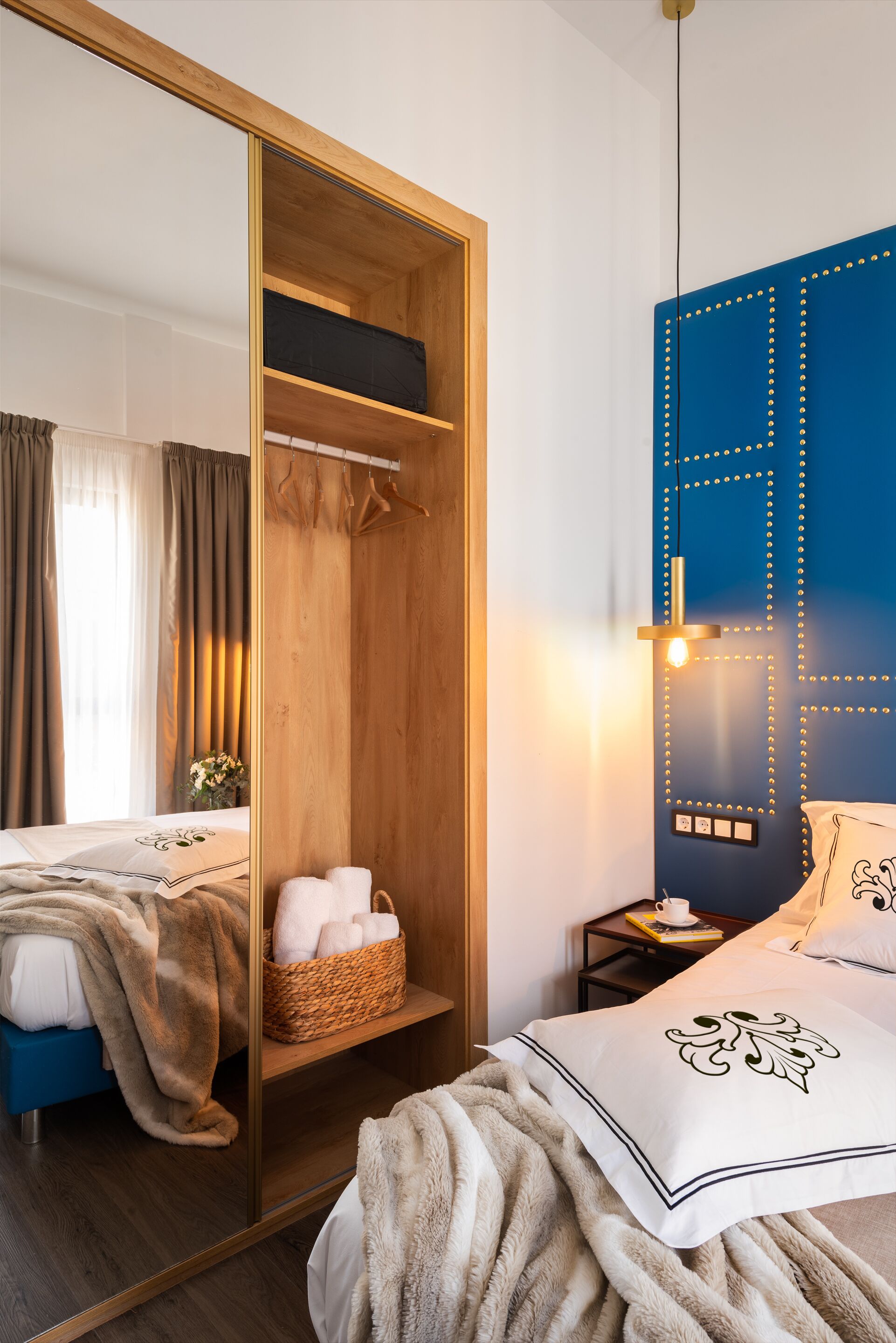A hotel room with a wooden wardrobe, a blue accent wall, a bedside table with a lamp, and a bed with a white pillow and blanket.
