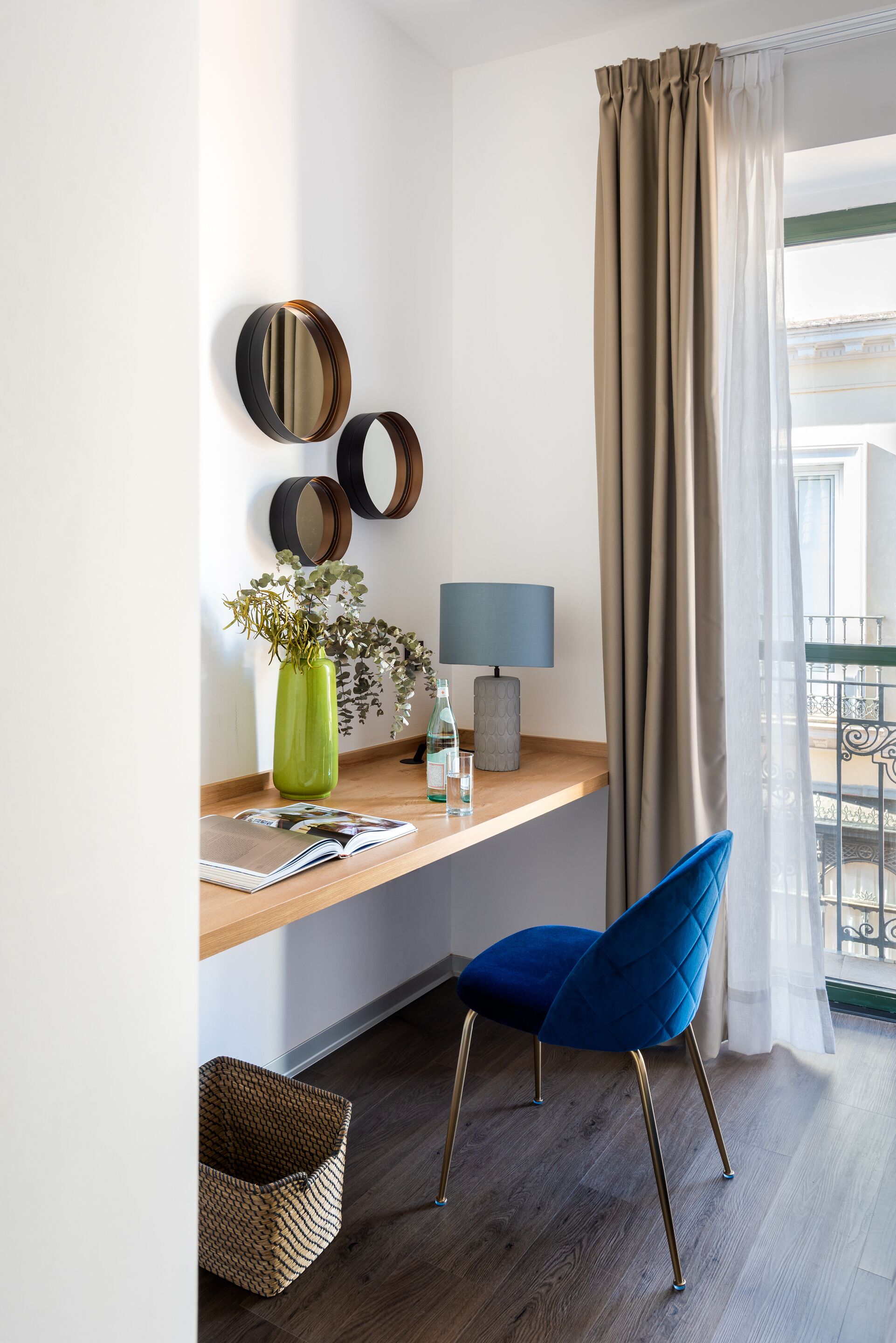 Habitació moderna amb escriptori de fusta, cadira blava, gerra verda amb plantes i finestra amb cortines.