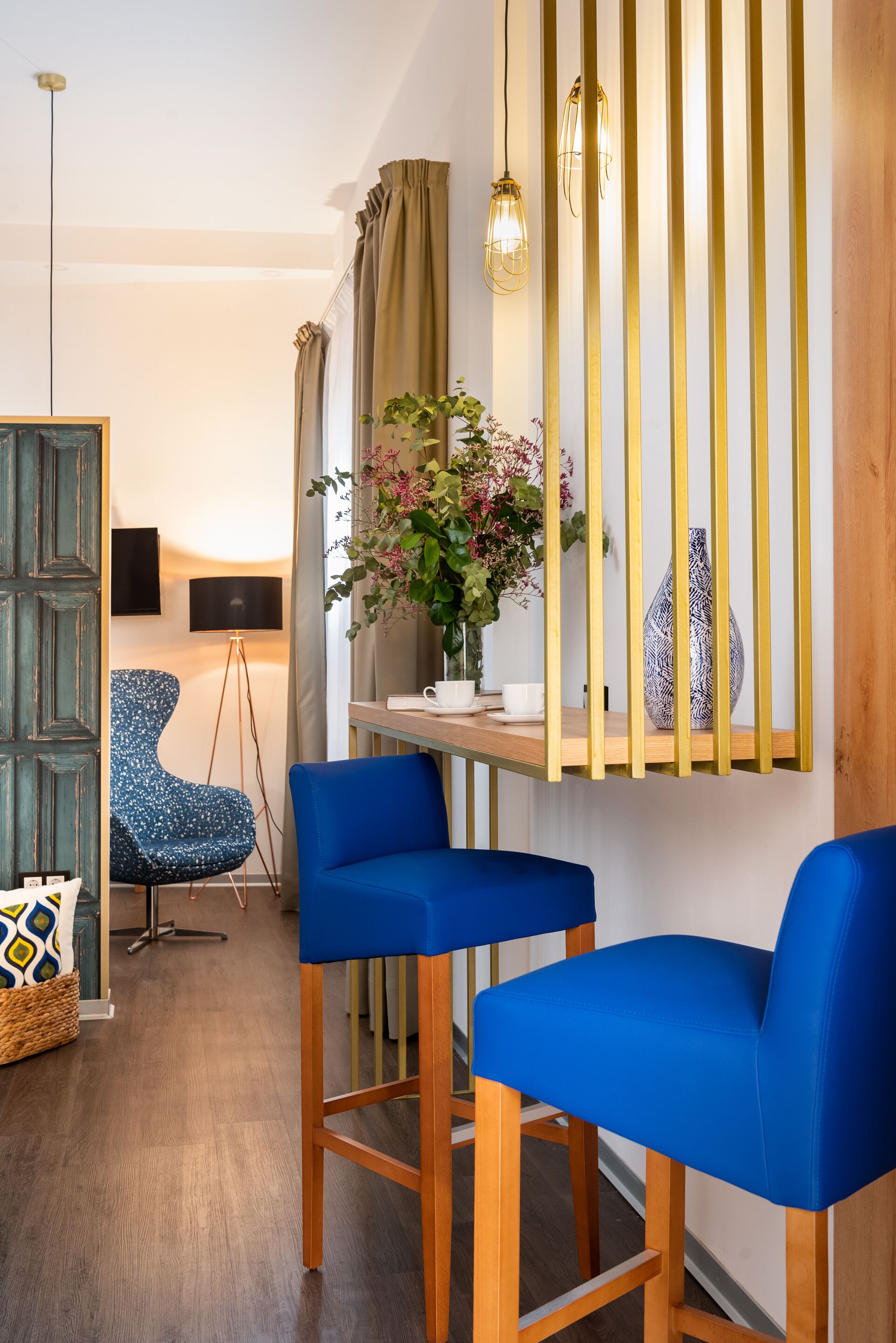 Una sala de estar moderna con suelo de madera, dos sillas azules, una mesita, un jarrón con flores, una lámpara y un televisor sobre un mueble bajo.