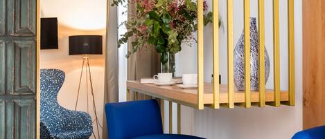 Una sala de estar moderna con suelo de madera, dos sillas azules, una mesita, un jarrón con flores, una lámpara y un televisor sobre un mueble bajo.