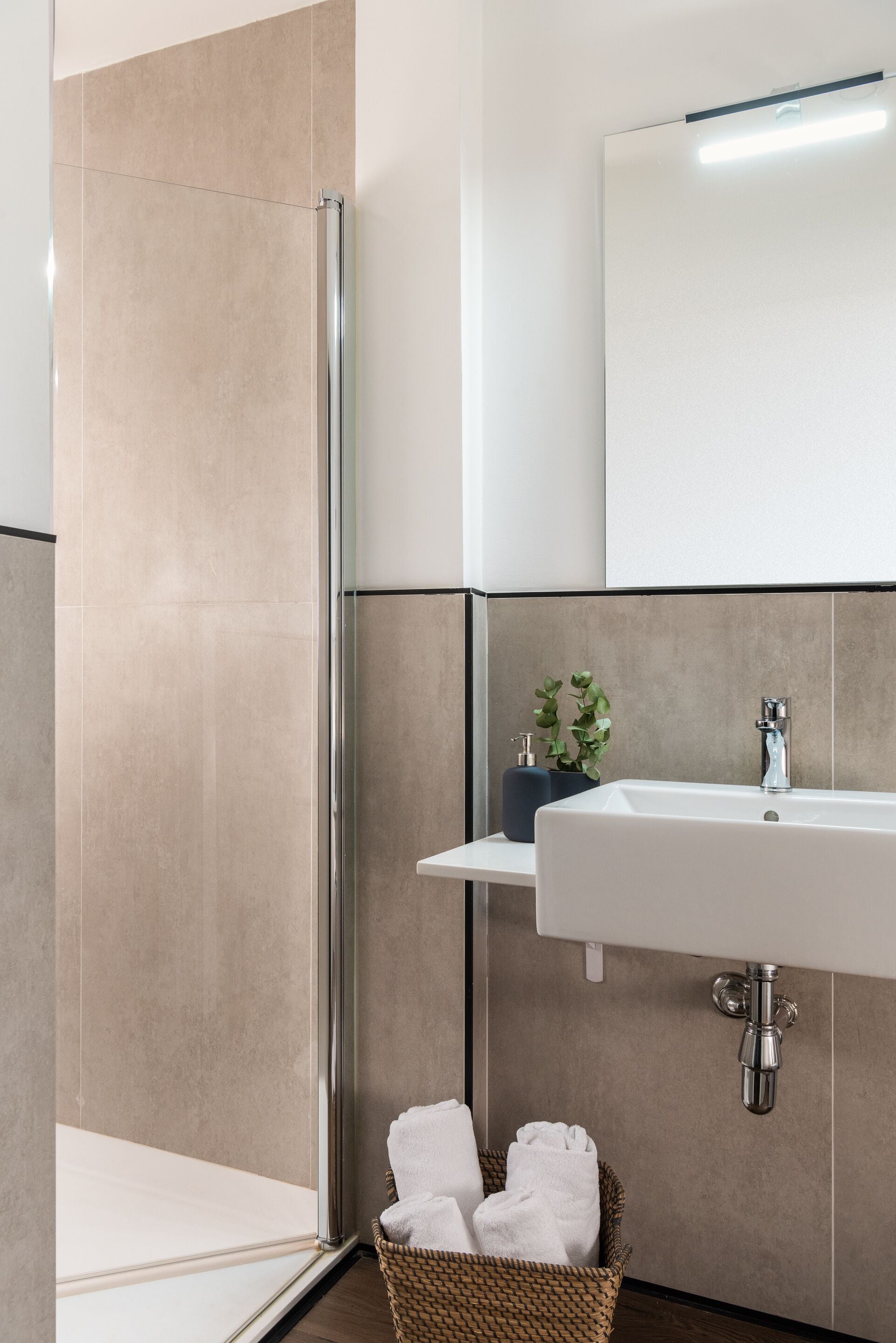 Un baño moderno con cabina de ducha de vidrio, un lavamanos blanco con grifo cromado y una cesta de mimbre con toallas.