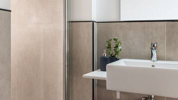 Un baño moderno con cabina de ducha de vidrio, un lavamanos blanco con grifo cromado y una cesta de mimbre con toallas.