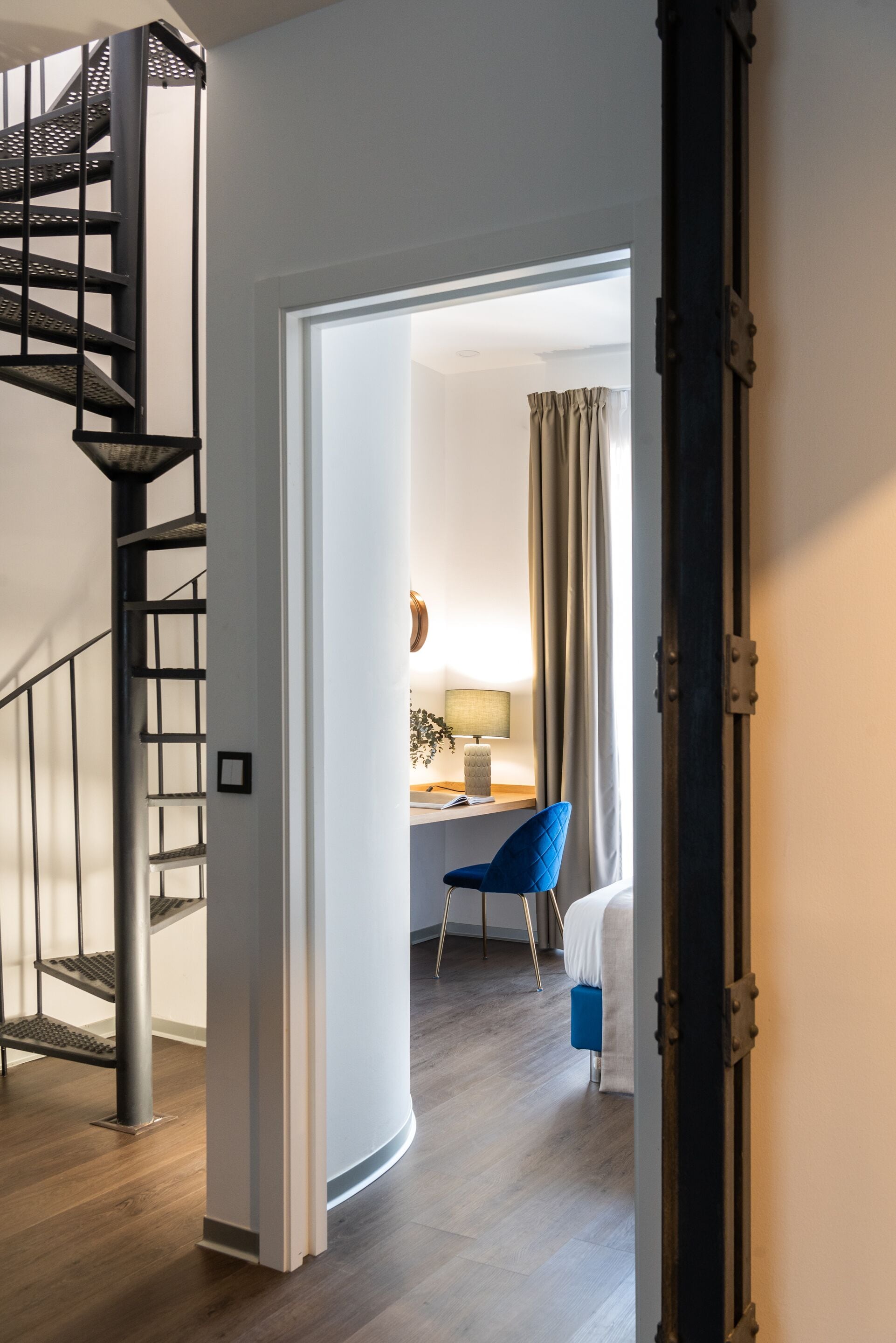 Habitación de hotel moderna con una escalera espiral, un escritorio con lámpara, una silla azul y una cama.