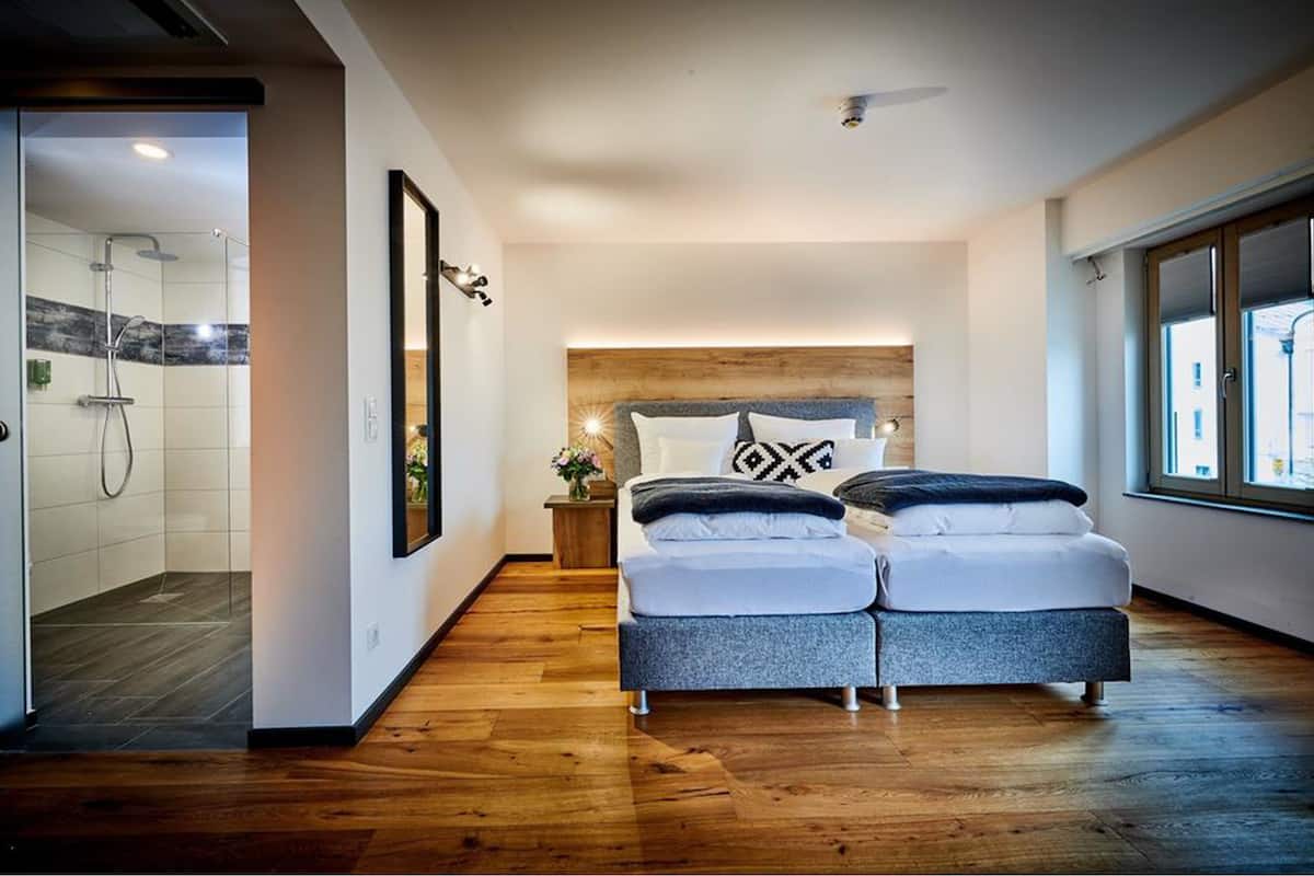 Un dormitorio moderno con cabecera de madera, cama doble con almohadones estampados, mesita de noche con un florero y un ventanal que deja entrar luz natural.