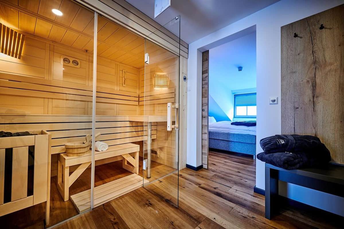 Un cuarto de sauna con bancos de madera y un mamparo de vidrio, que da acceso a un dormitorio con cama y mesita de noche.