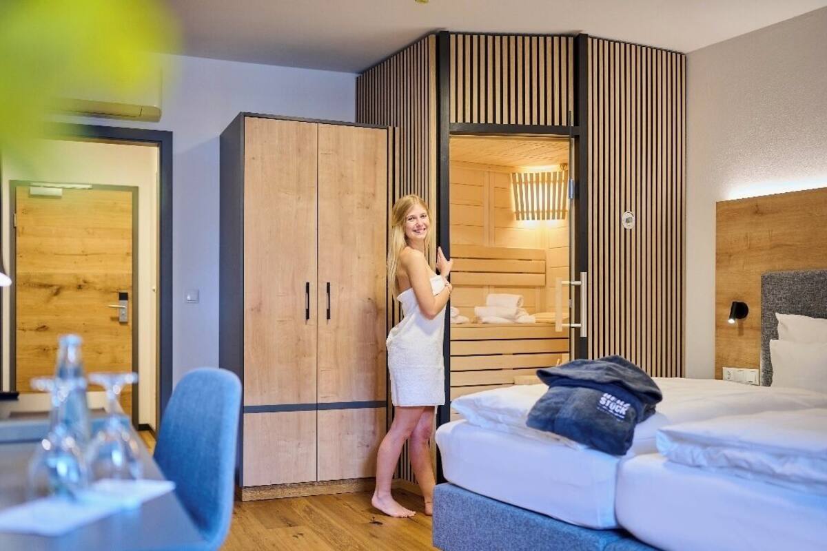 Una habitación de hotel con un armario de madera, una cama con almohada y una mesita con un objeto de vidrio.