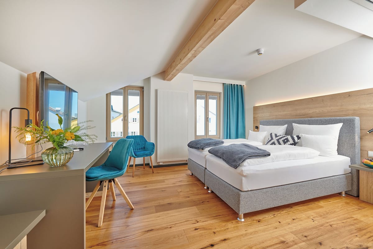Una habitación de hotel moderna con techo de madera, una cama grande con ropa de cama gris, un escritorio con una silla y vistas al exterior a través de amplios ventanales.