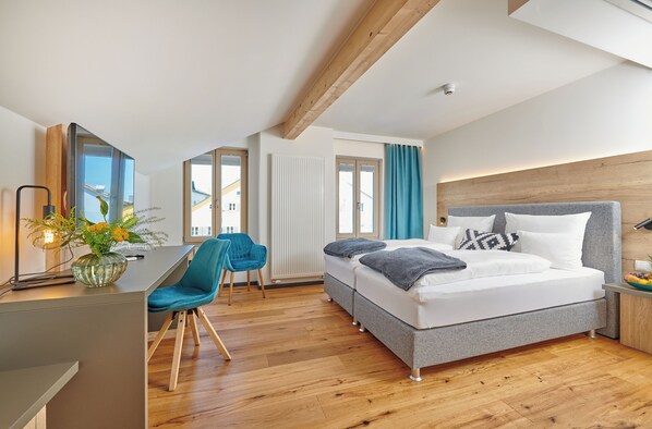 Una habitación de hotel moderna con techo de madera, una cama grande con ropa de cama gris, un escritorio con una silla y vistas al exterior a través de amplios ventanales.