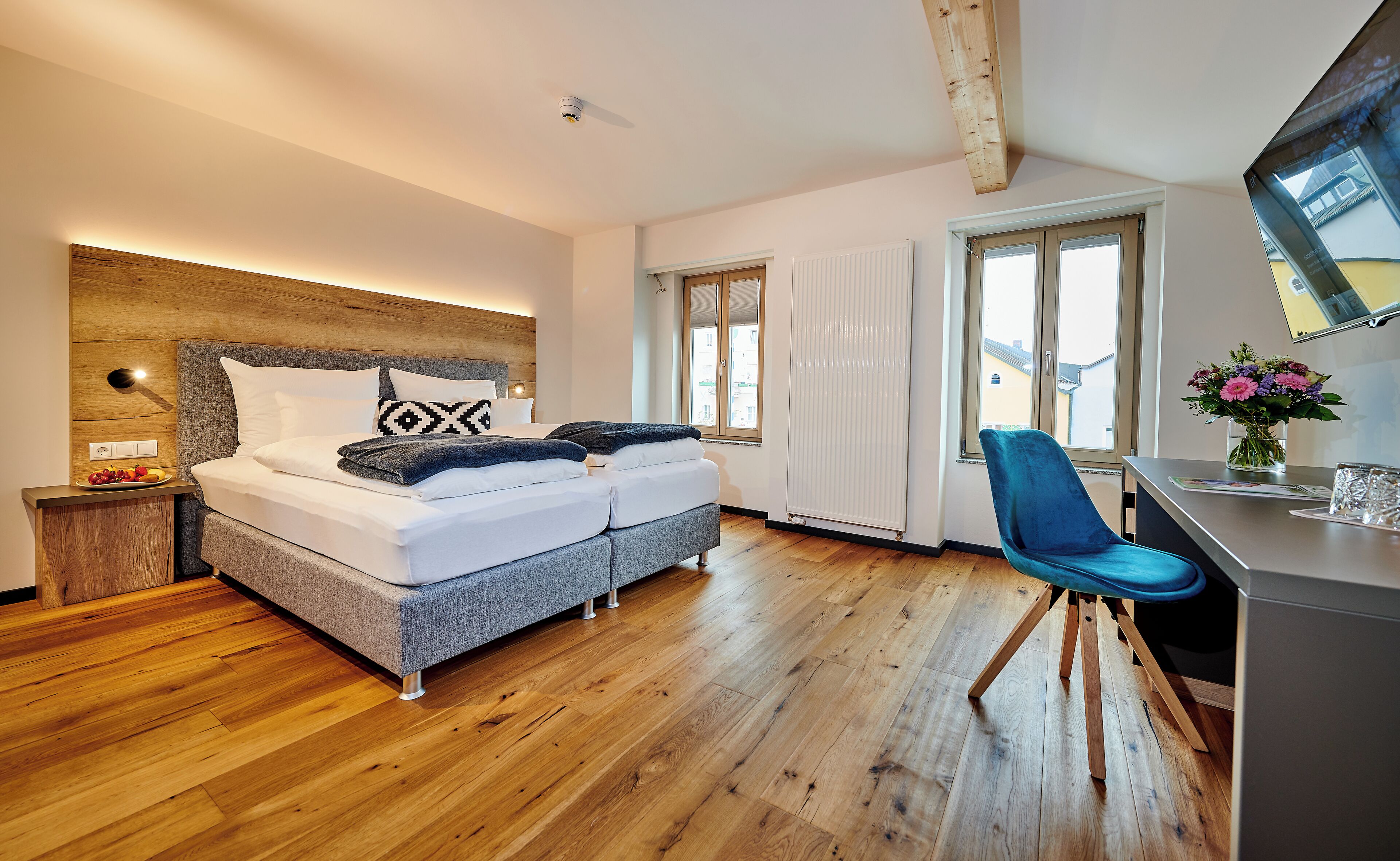 A modern bedroom with a wooden headboard, a bed with white bedding, a desk with a chair, and a window with a view of buildings.