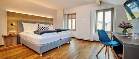 A modern bedroom with a wooden headboard, a bed with white bedding, a desk with a chair, and a window with a view of buildings.