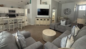 Ein modernes Wohnzimmer mit grauem Sofa, Fernseher und Küchenzeile mit weißen Schränken und einer Mikrowelle.