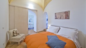 A bedroom with an orange bedspread, a chair with a bag, a painting on the wall, and a door leading to another room. - Suite Malu' (Praiano)