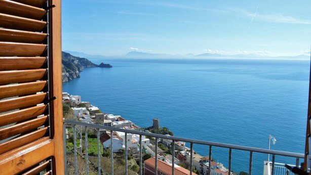 A coastal view from a balcony with a railing, overlooking a calm sea and a small town with buildings and a mountain in the distance. - Suite Malu' (Praiano)