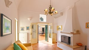 A spacious living room with a sofa, a fireplace, and a dining area visible through an open door. - Casa Alessia (Praiano)