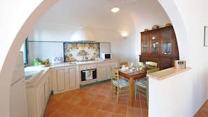 A kitchen with white cabinets, a tiled backsplash, a dining table with chairs, and a wooden cabinet with glass doors. - Casa Alessia (Praiano)