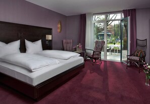 Ein Hotelzimmer mit einem großen Bett, zwei Sesseln, einem Tisch und Blick auf den Garten durch eine Glastür.