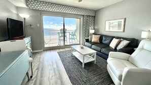 A living room with a sofa, coffee table, and a TV. There is a balcony with a view of the beach. - Royal Palms 402 1 Bedroom Condo by RedAwning (Gulf Shores)