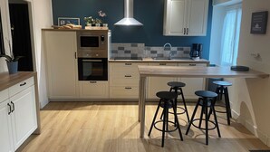 Cocina moderna con armarios blancos, horno empotrado, microondas y cafetera. Hay tres taburetes negros en la isla.