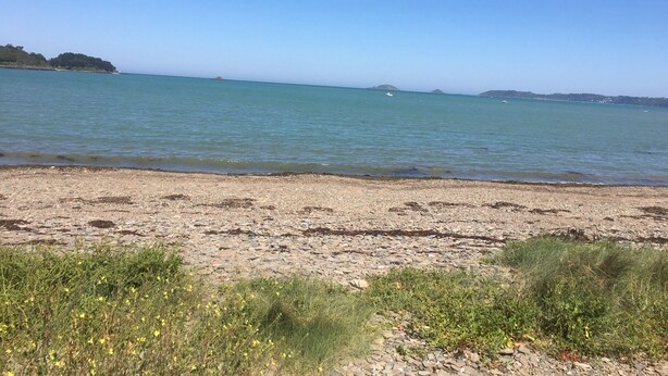 Una playa con agua azul cristalina, costa rocosa y colinas al fondo.