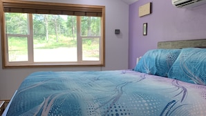 Un dormitorio con una cama grande, una ventana con vistas a árboles, un ventilador de techo y un equipo de aire acondicionado.