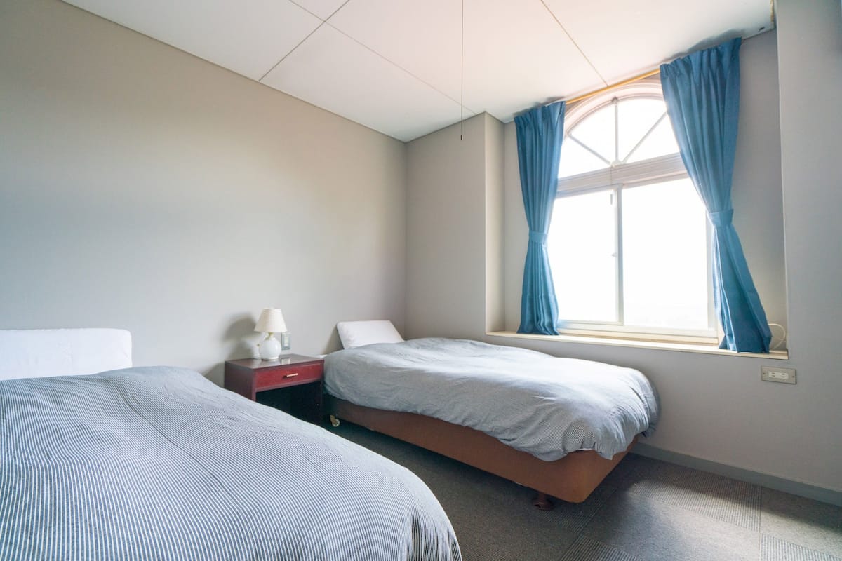 A room with two beds, a nightstand, a lamp, and a window with curtains.