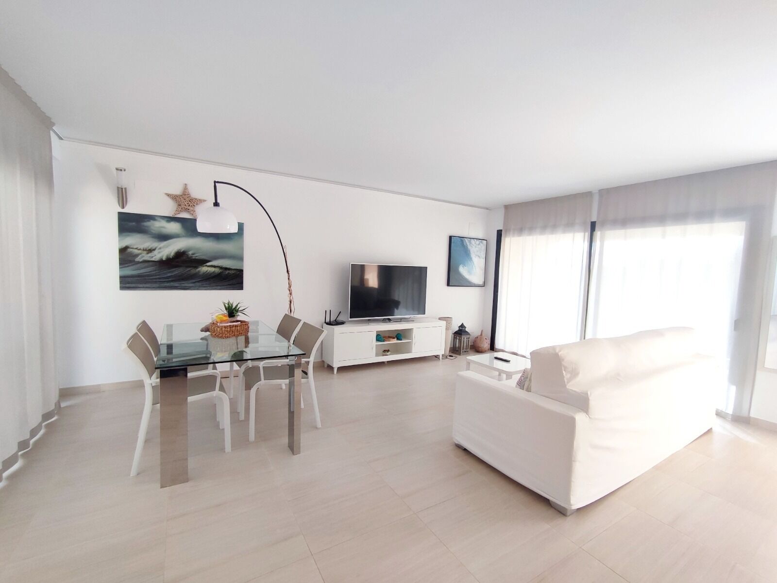 A modern living room with a glass dining table, white sofa, flat-screen TV, and artwork on the walls.