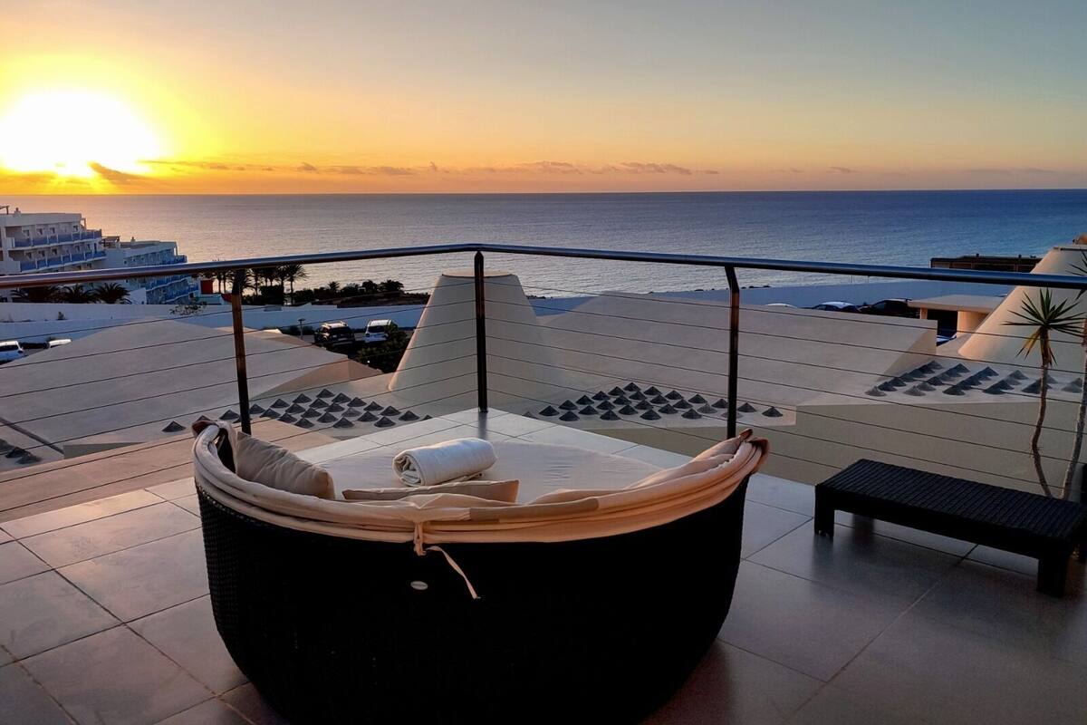 A balcony with a cushioned seating area and a view of the sunset over the sea.