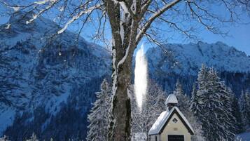 Un paysage enneigé avec un grand arbre, un petit bâtiment doté d’un toit en pente et, au loin, des montagnes recouvertes de neige.
