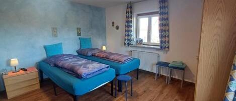 A bedroom with two beds, a wooden floor, a window with curtains, a radiator, and a small table.