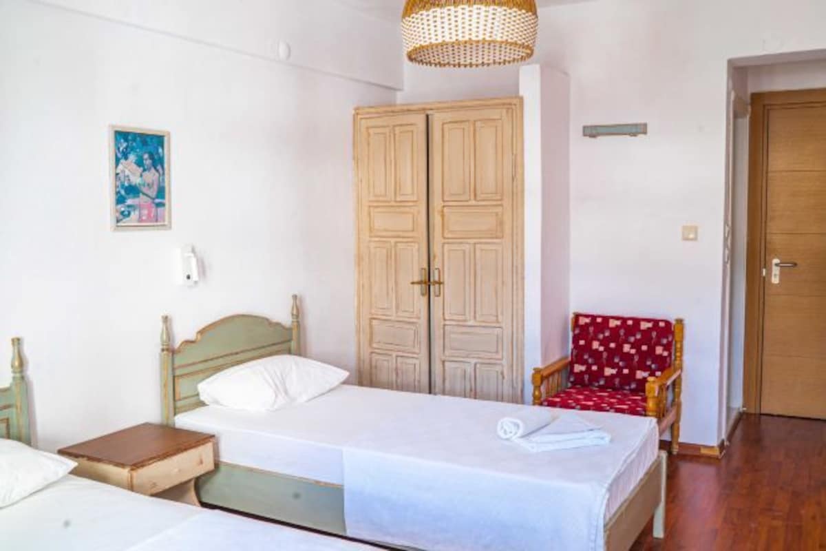 Una habitación con dos camas individuales, un armario de madera, una silla roja con estampado y una puerta.