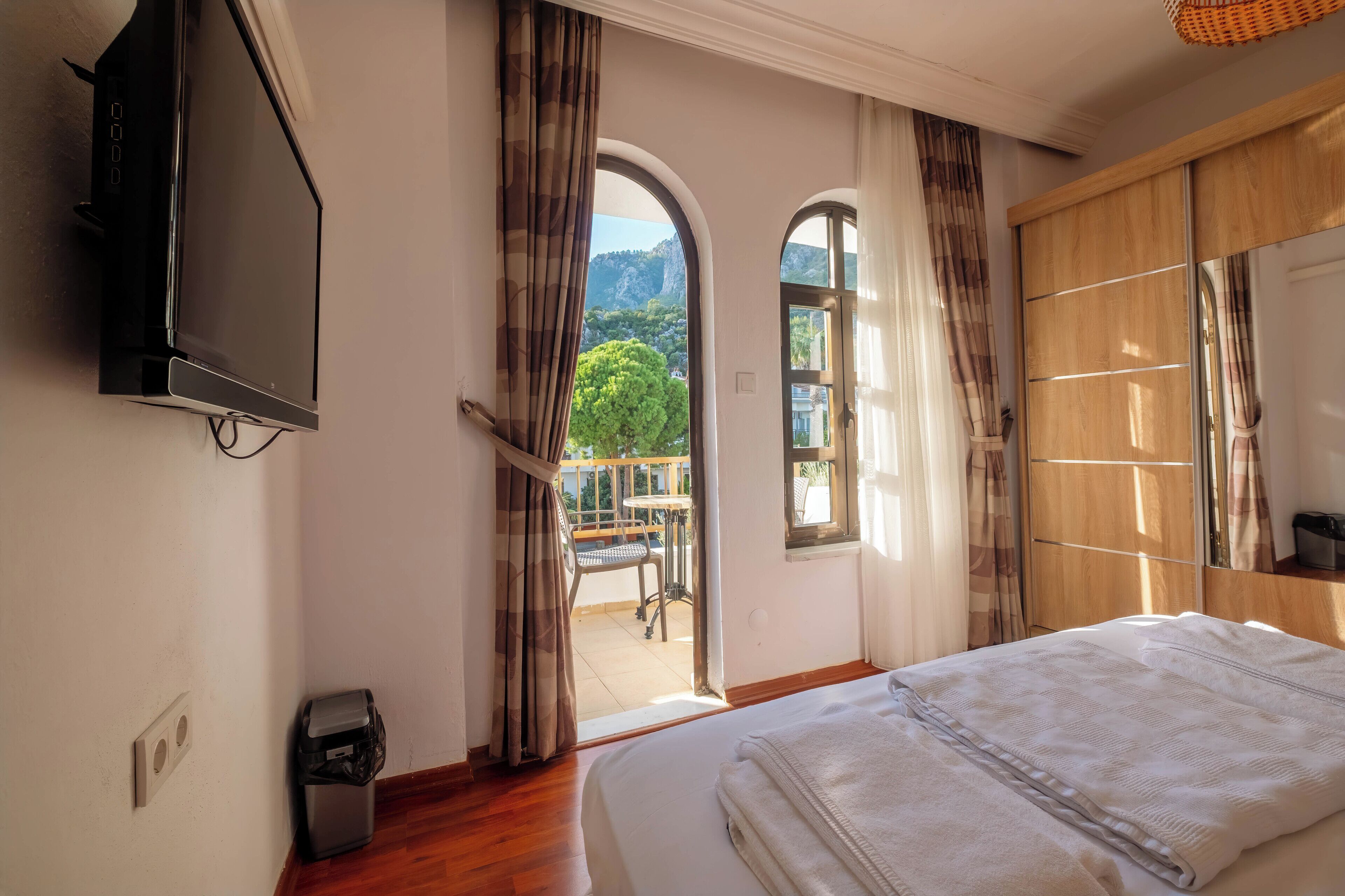 A bedroom with a bed, a TV, and a balcony with a view of greenery and mountains.