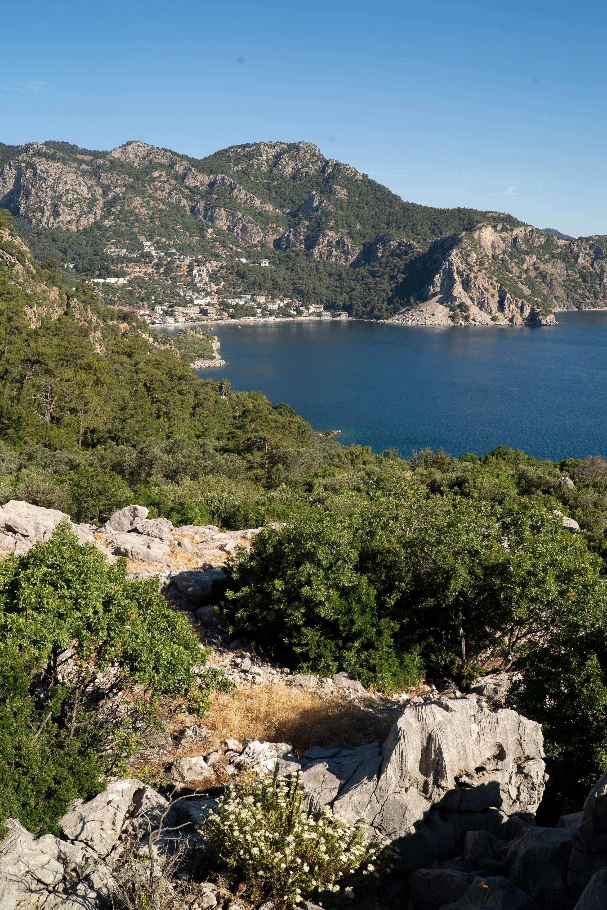 A mountainous landscape with a clear view of a coastal area, rocky terrain, and dense greenery.