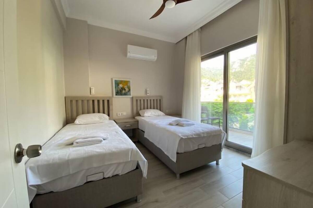 Une chambre d’hôtel avec deux lits simples, un ventilateur de plafond, un appareil de climatisation, une fenêtre donnant sur un espace verdoyant et une petite table.