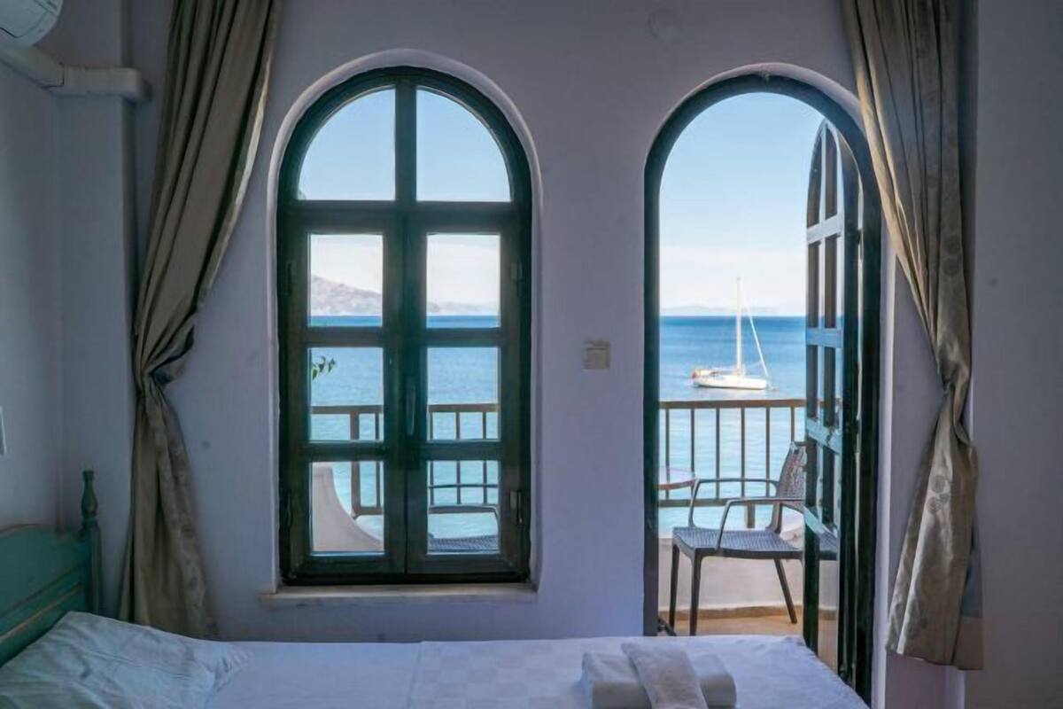 Une chambre avec un lit, deux fenêtres avec des rideaux, une vue sur la mer et une chaise à l’extérieur.