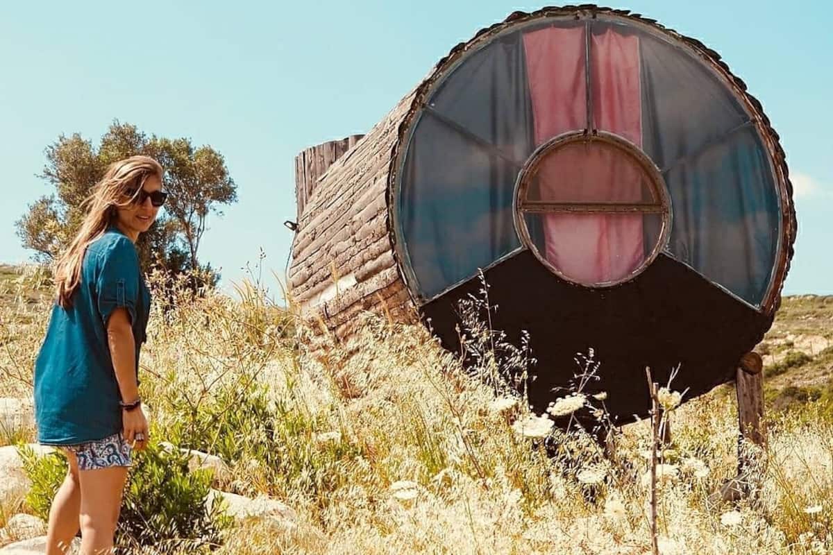 A person standing next to a large, cylindrical structure with a circular window, surrounded by dry grass and a clear sky.