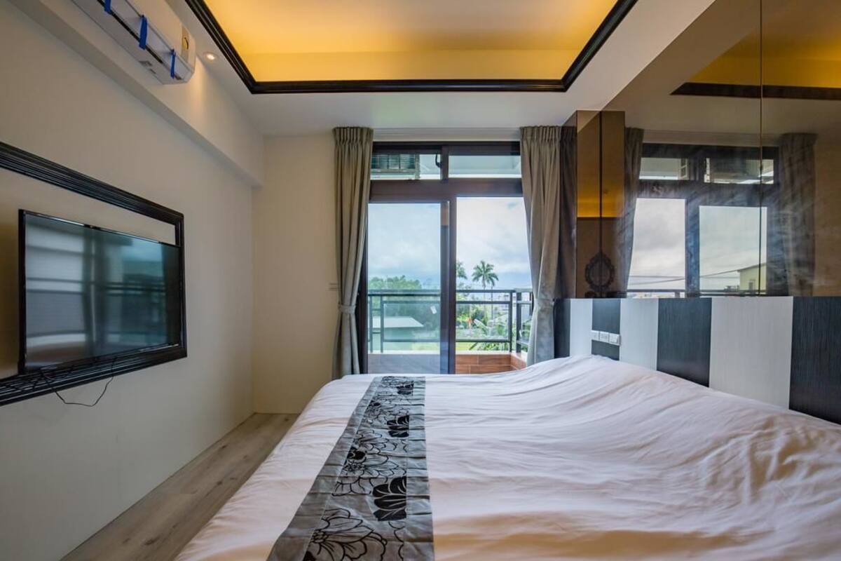 A bedroom with a large bed, a TV mounted on the wall, and a balcony with a view of the outdoors.