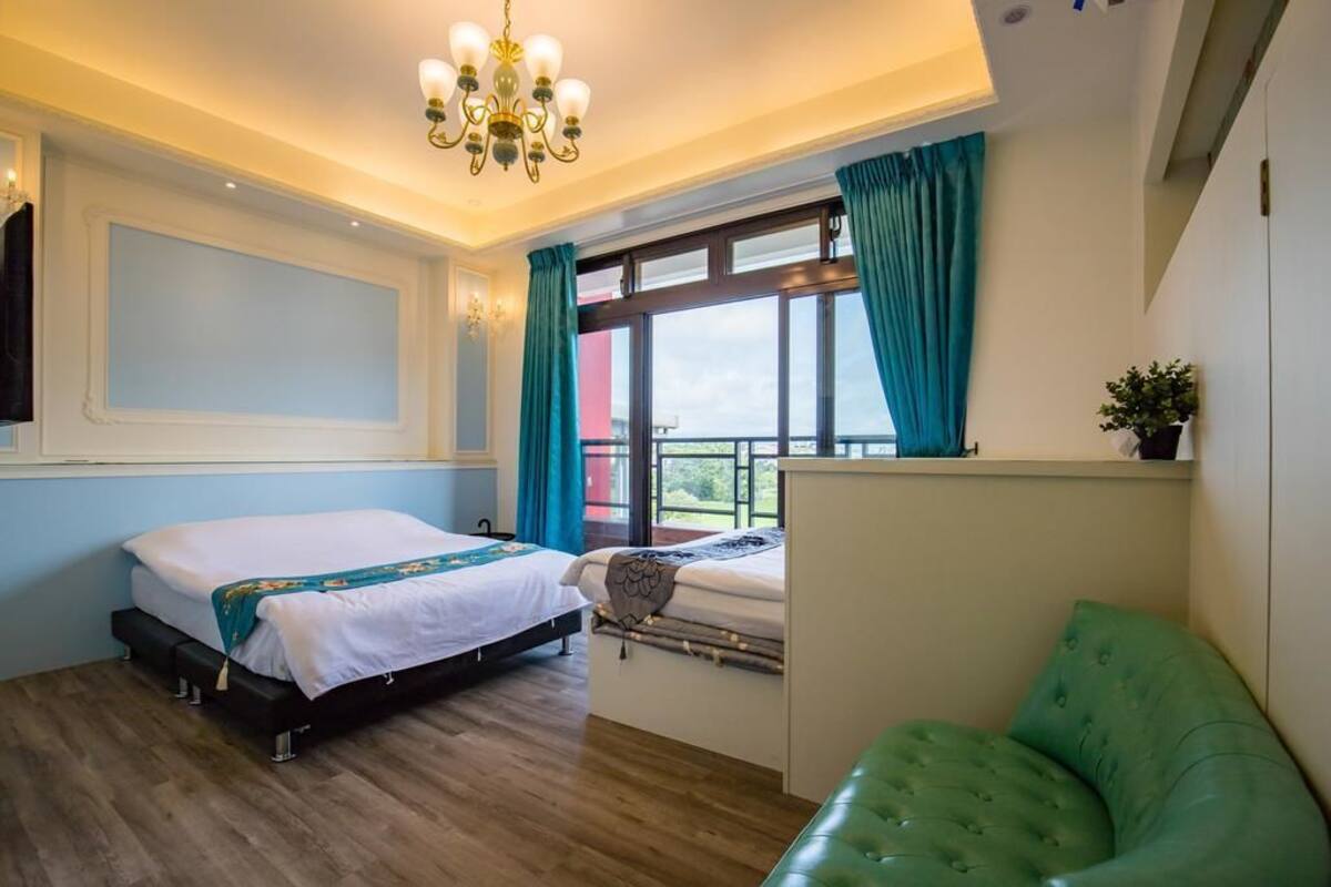A bedroom with a bed, a green sofa, a window with curtains, a chandelier, and a view of the outdoors.