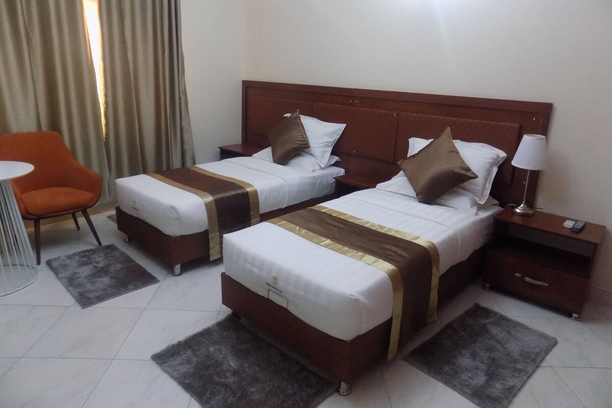 A hotel room with two beds, a nightstand, a lamp, a chair, and a small table.