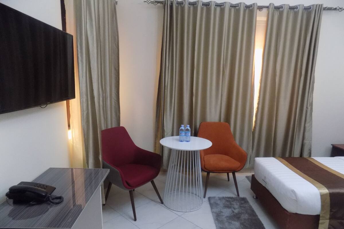 A hotel room with a bed, two chairs, a small table with water bottles, a desk with a phone, and curtains.