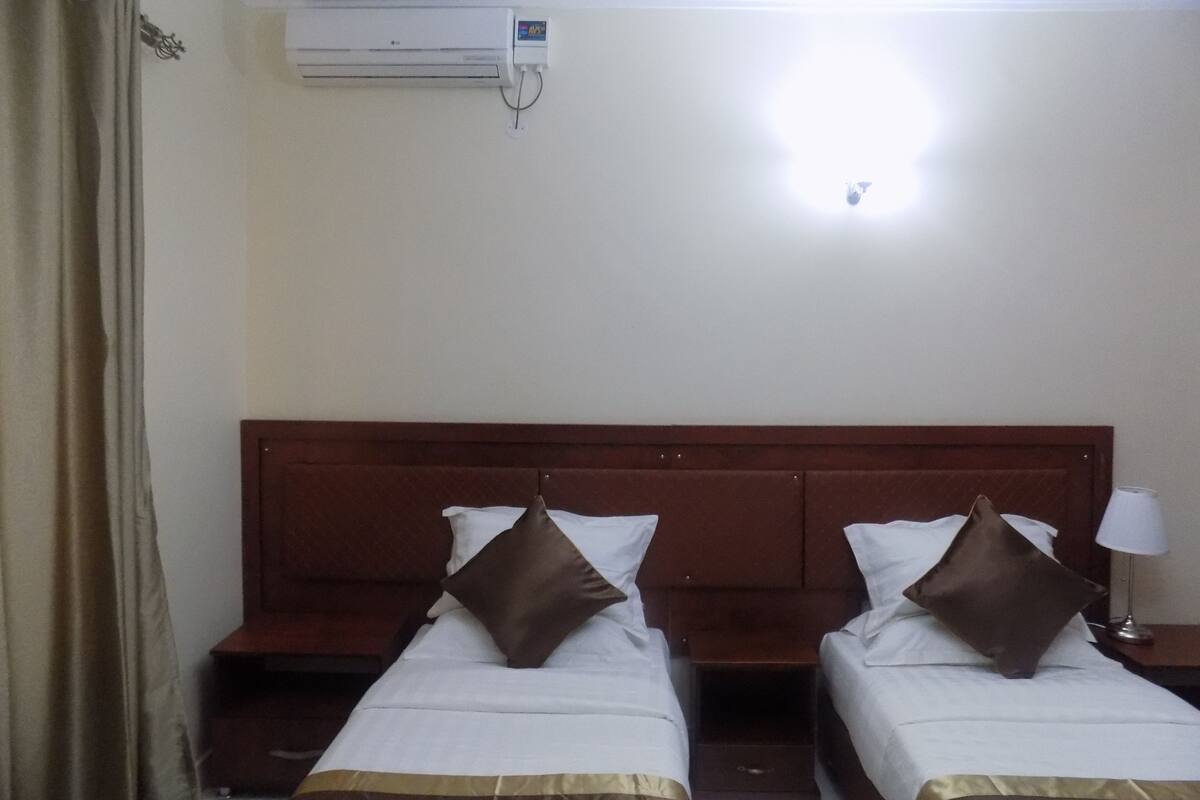 A hotel room with two single beds, a wooden headboard, a nightstand with a lamp, and an air conditioning unit on the wall.