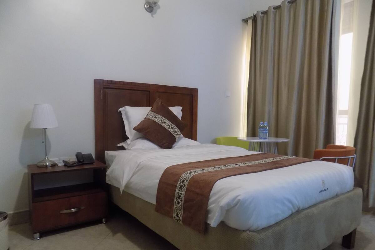 A hotel room with a bed, bedside table, lamp, and a small table with water bottles.