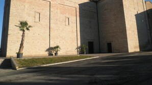 A large, rectangular building with a flat roof and a symmetrical facade. There are two small windows on the second floor and a door on the ground floor. The building is made of light-colored stone or brick, and there is a palm tree and two potted plants in front of it. - B&B Severini (Morrovalle)