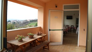 A balcony with a wooden table and potted plants, a door leading to a dining area with tables and chairs, and a view of a landscape with buildings in the distance. - B&B Severini (Morrovalle)