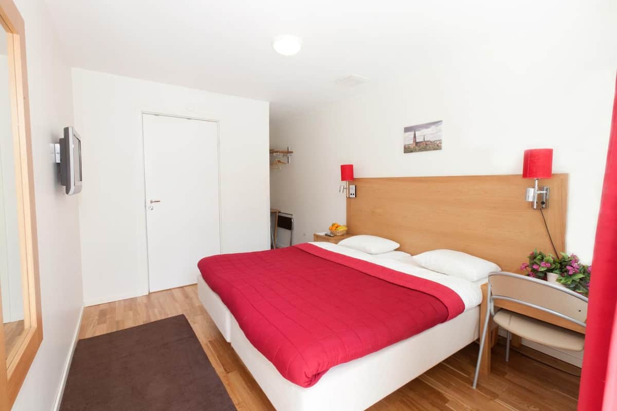 A hotel room with a bed, a wooden headboard, a bedside table with a lamp, a chair, and a small table with a basket of fruit.