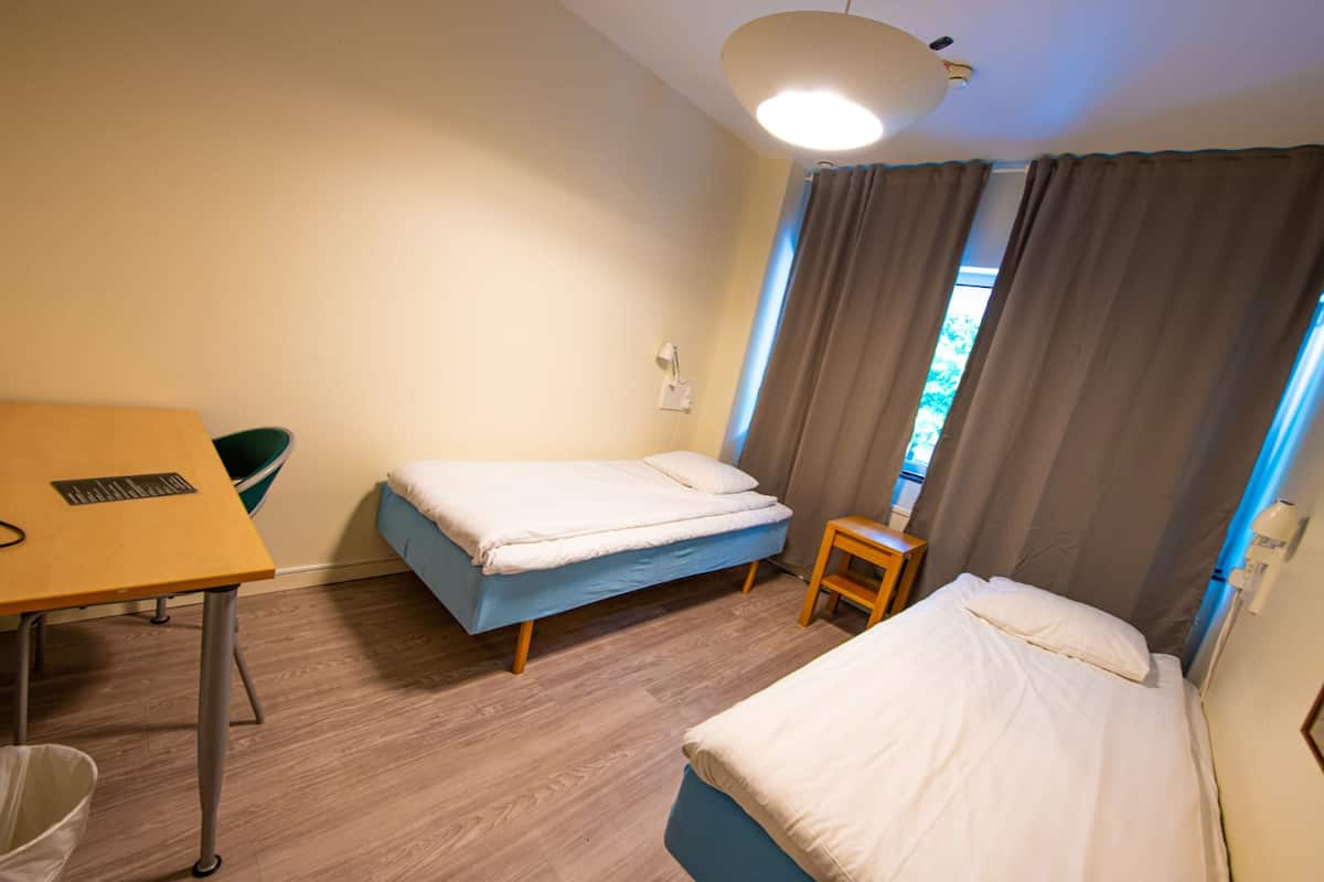 A small, clean room with a wooden floor, two single beds, a small table, and a chair.